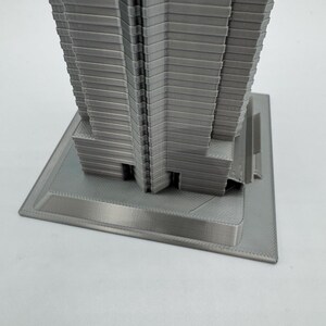 May include: A gray, 3D-printed model of a skyscraper. The building has a rectangular base and a multi-tiered design, with horizontal lines creating a textured facade. The model is made of a light gray material.