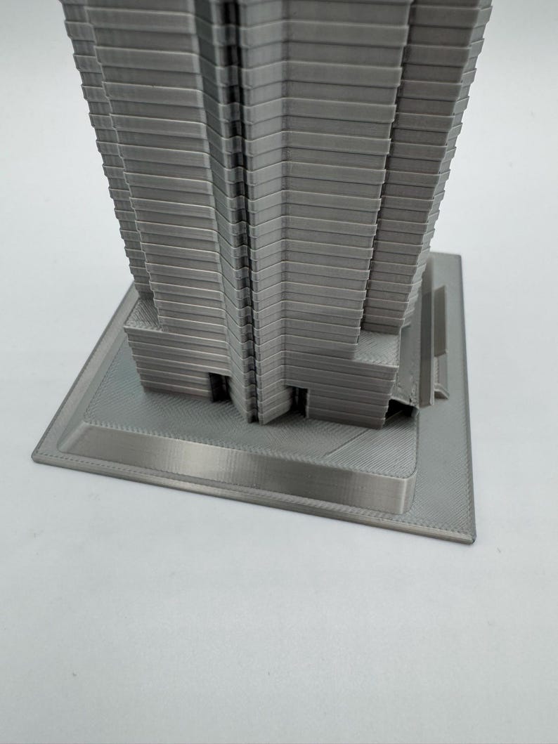 May include: A gray, 3D-printed model of a skyscraper. The building has a rectangular base and a multi-tiered design, with horizontal lines creating a textured facade. The model is made of a light gray material.