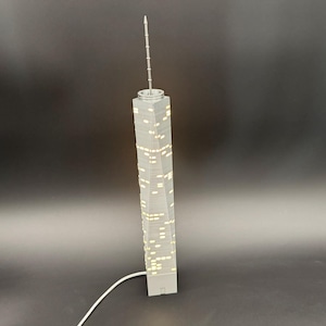 May include: A gray model of a skyscraper with illuminated windows and a tall antenna. The building is rectangular with a slightly twisted design. A white cord extends from the base.