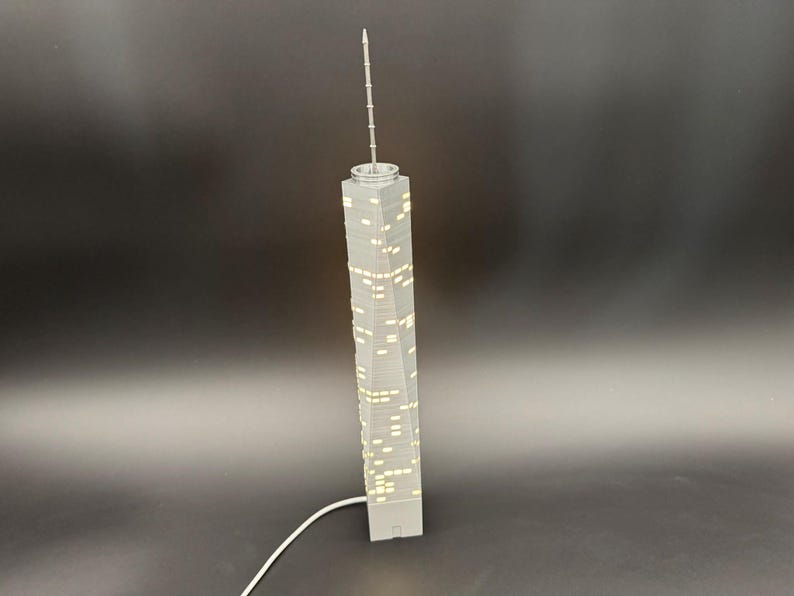 May include: A gray model of a skyscraper with illuminated windows and a tall antenna. The building is rectangular with a slightly twisted design. A white cord extends from the base.