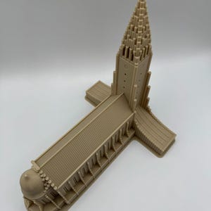 May include: A tan-colored architectural model of a church with a tall, pointed steeple and a cross at the top. The model has a long rectangular main building with a domed structure at one end and a cross-shaped base.