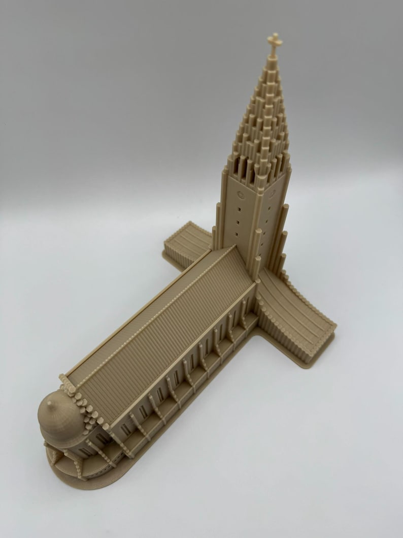 May include: A tan-colored architectural model of a church with a tall, pointed steeple and a cross at the top. The model has a long rectangular main building with a domed structure at one end and a cross-shaped base.