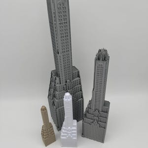May include: A collection of gray and beige miniature skyscraper models, varying in size and detail. The models showcase architectural features, with a wooden ruler in the foreground for scale. The buildings are arranged on a white surface.