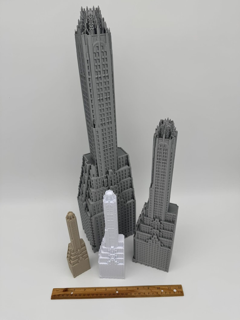 May include: A collection of gray and beige miniature skyscraper models, varying in size and detail. The models showcase architectural features, with a wooden ruler in the foreground for scale. The buildings are arranged on a white surface.