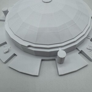 May include: A light gray architectural model featuring a domed roof with layered details and a circular top. The model includes surrounding rectangular platforms and cylindrical structures, all in a monochromatic color scheme.