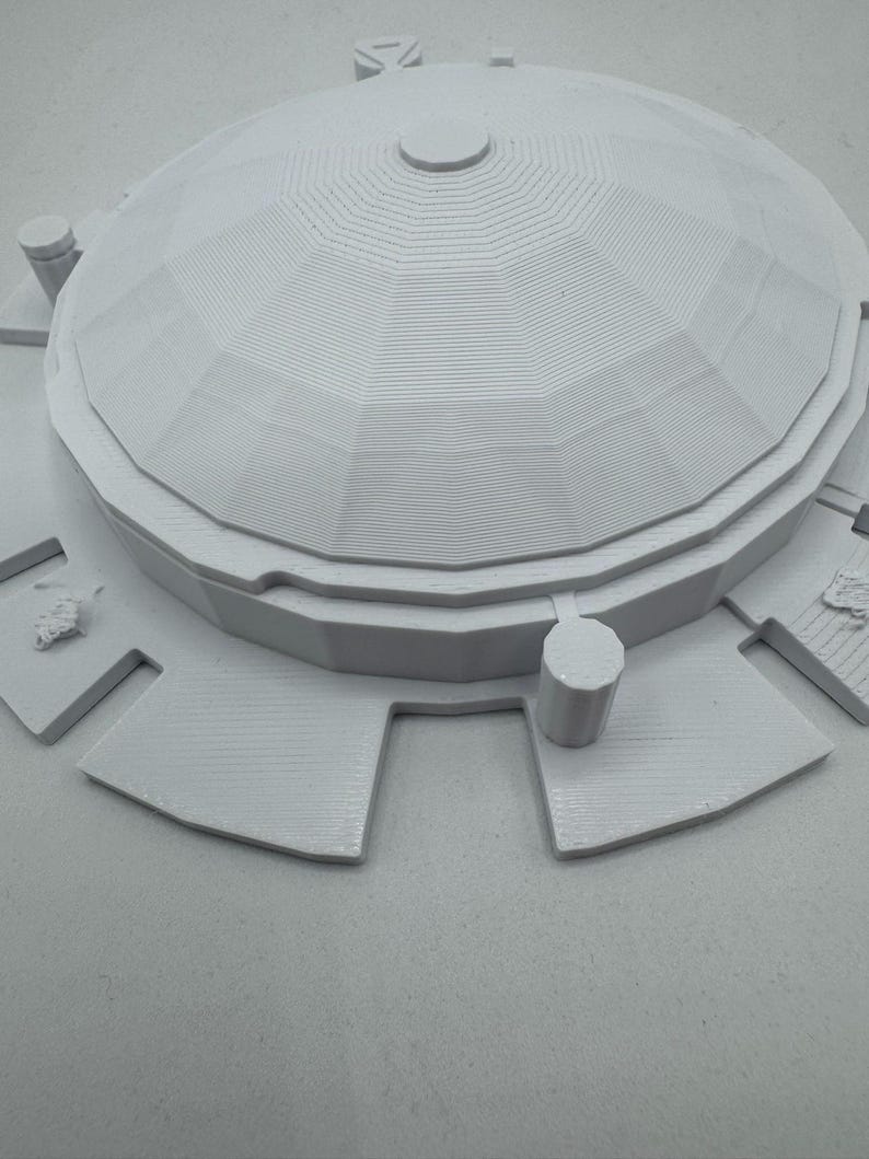 May include: A light gray architectural model featuring a domed roof with layered details and a circular top. The model includes surrounding rectangular platforms and cylindrical structures, all in a monochromatic color scheme.