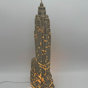 May include: A beige, multi-tiered skyscraper lamp featuring a detailed architectural design with a central spire. The structure is illuminated from within, casting a warm, glowing light through numerous small, rectangular window cutouts.