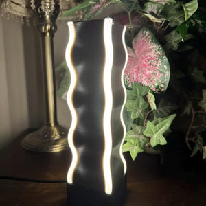 K&ouml;nnte beinhalten: Eine schwarze Lampe mit wellenf&ouml;rmigen Kanten und vertikalen wei&szlig;en Lichtstreifen. Die Lampe hat einen rechteckigen Sockel und eine hohe, zylindrische Form. Die Lampe steht auf einer dunklen Holzoberfl&auml;che, mit einer traditionellen Lampe und Gr&uuml;nzeug im Hintergrund.