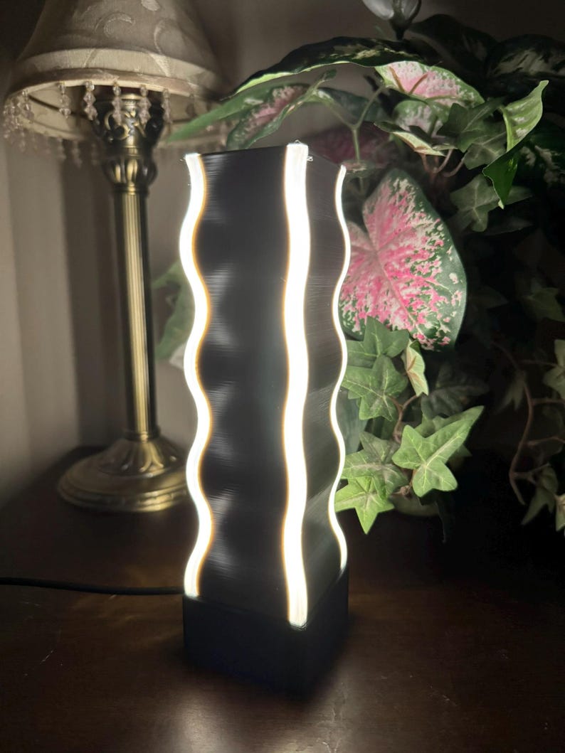 K&ouml;nnte beinhalten: Eine schwarze Lampe mit wellenf&ouml;rmigen Kanten und vertikalen wei&szlig;en Lichtstreifen. Die Lampe hat einen rechteckigen Sockel und eine hohe, zylindrische Form. Die Lampe steht auf einer dunklen Holzoberfl&auml;che, mit einer traditionellen Lampe und Gr&uuml;nzeug im Hintergrund.