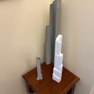 May include: Three miniature skyscraper models in gray and white are displayed on a small, dark wooden table. A ruler is placed in front of the models. The tallest model is approximately 10 inches tall.
