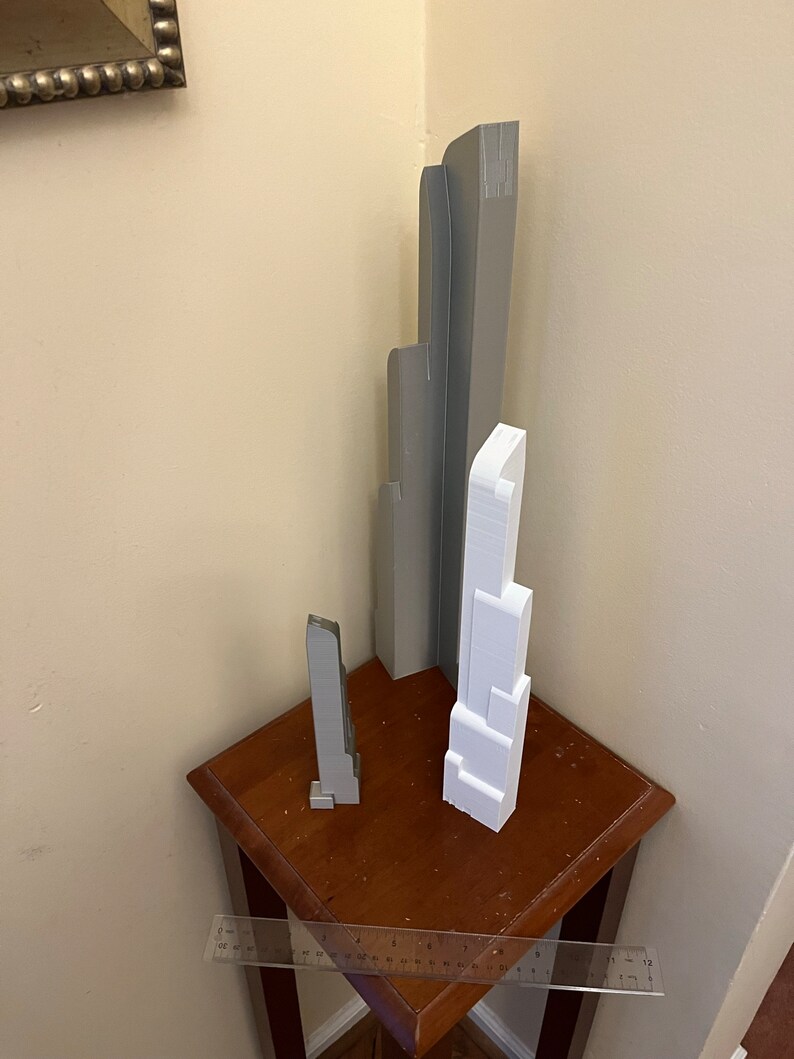 May include: Three miniature skyscraper models in gray and white are displayed on a small, dark wooden table. A ruler is placed in front of the models. The tallest model is approximately 10 inches tall.
