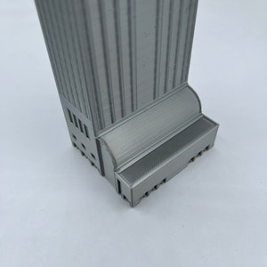 May include: A gray 3D printed model of a tall building with a detailed facade and a base with a textured surface.