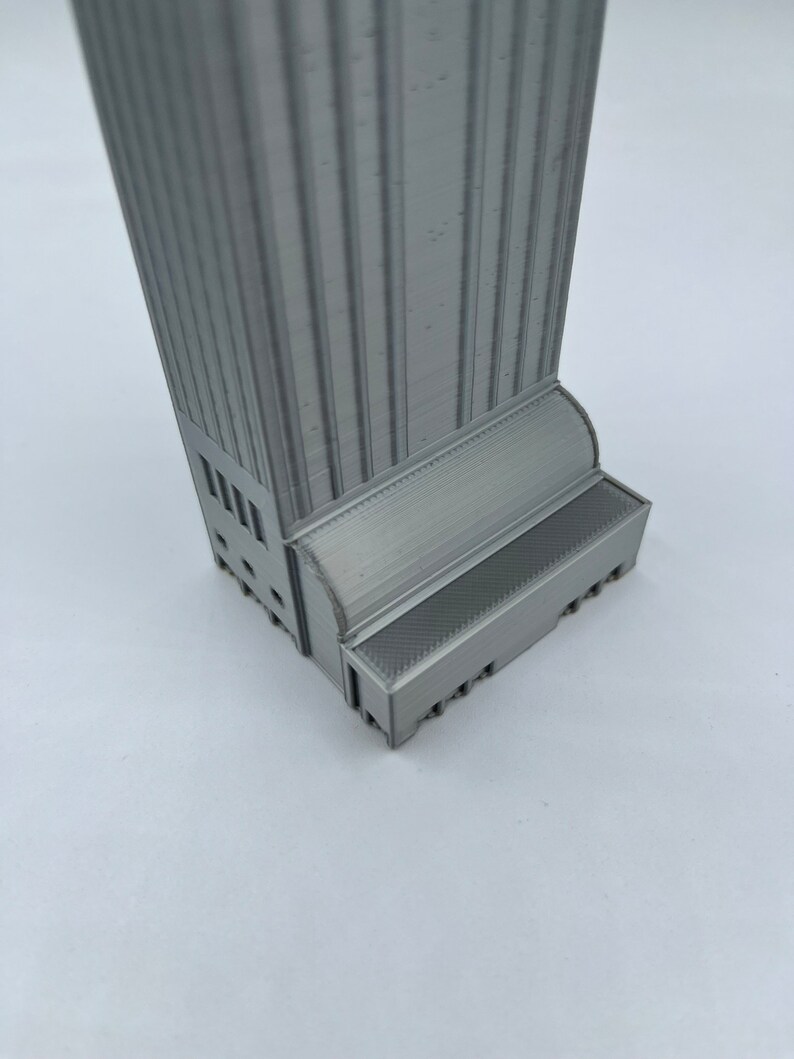 May include: A gray 3D printed model of a tall building with a detailed facade and a base with a textured surface.