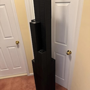 May include: A black architectural model of a skyscraper. The model has a rectangular base and multiple levels, with a textured facade. The building is positioned next to a white door and a beige wall.
