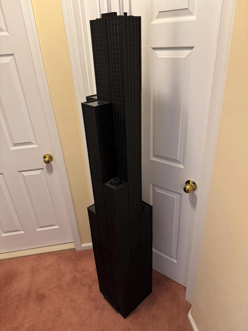May include: A black architectural model of a skyscraper. The model has a rectangular base and multiple levels, with a textured facade. The building is positioned next to a white door and a beige wall.