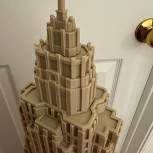 Może przedstawiać: Szczeg&oacute;łowy model 3D wydrukowany w kolorze beżowym, przedstawiający wieżowiec z misternymi detalami architektonicznymi. Model został zaprojektowany tak, aby przypominał nowoczesny wieżowiec z ostrym szczytem.