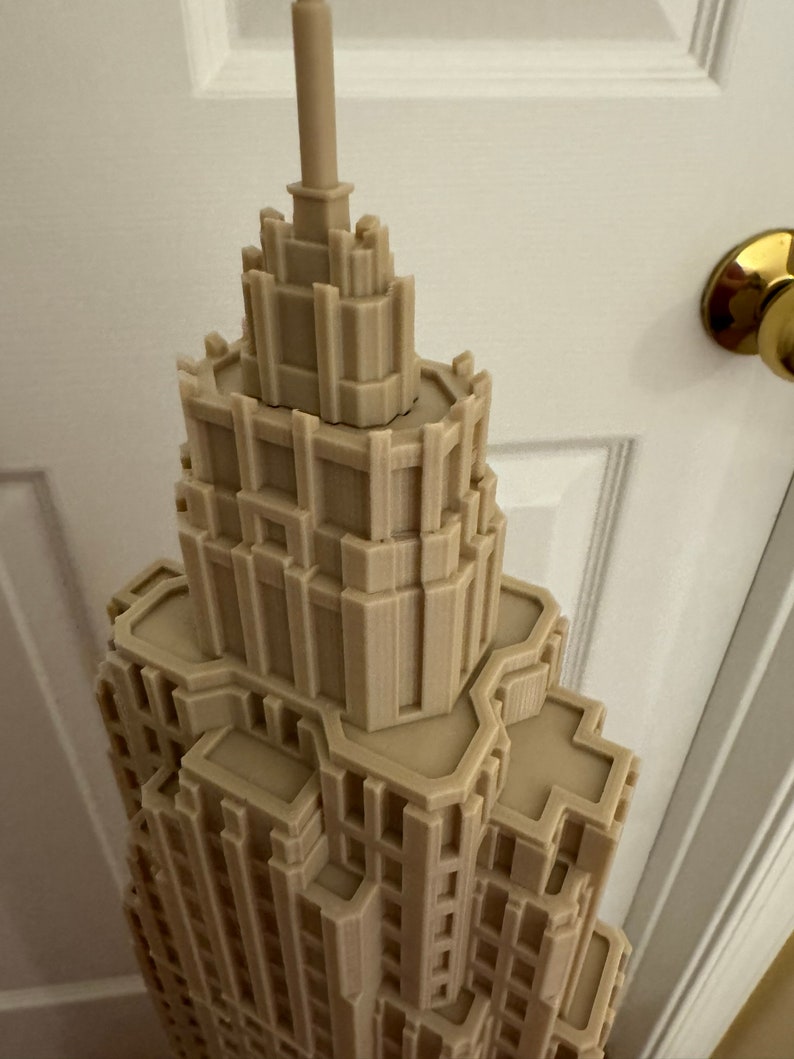 Może przedstawiać: Szczeg&oacute;łowy model 3D wydrukowany w kolorze beżowym, przedstawiający wieżowiec z misternymi detalami architektonicznymi. Model został zaprojektowany tak, aby przypominał nowoczesny wieżowiec z ostrym szczytem.