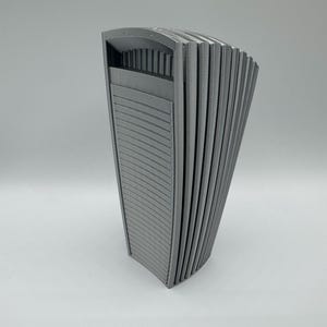 May include: A gray, vertical, fan-shaped object with a series of parallel, ridged panels. The top section has a rectangular opening with vertical supports. The object is made of a solid material and has a smooth, matte finish.