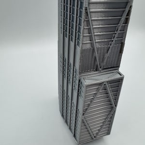 May include: A 3D printed architectural model of a skyscraper in a metallic silver finish. The structure features a rectangular base with a distinct protruding section, showcasing intricate geometric grid patterns and vertical lines on its exterior.