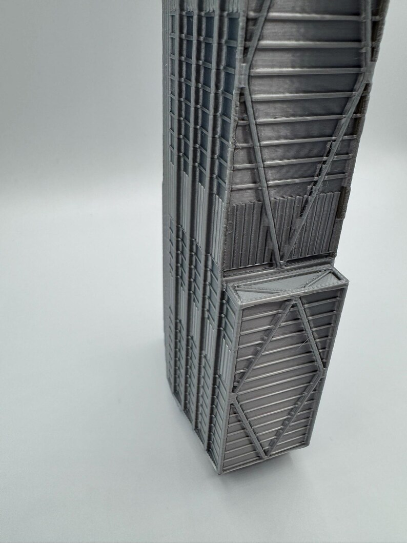 May include: A 3D printed architectural model of a skyscraper in a metallic silver finish. The structure features a rectangular base with a distinct protruding section, showcasing intricate geometric grid patterns and vertical lines on its exterior.