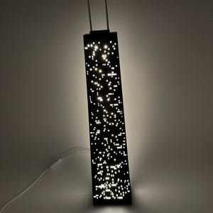 May include: A black rectangular pendant light fixture with numerous small, bright white lights. The light hangs from two thin, white cords and has a white power cord. The fixture is illuminated, casting a soft glow on the surrounding wall.