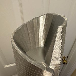 May include: A silver 3D printed model of a tall, cylindrical building with a unique, angled design. The model is detailed with intricate lines and textures, showcasing the capabilities of 3D printing.