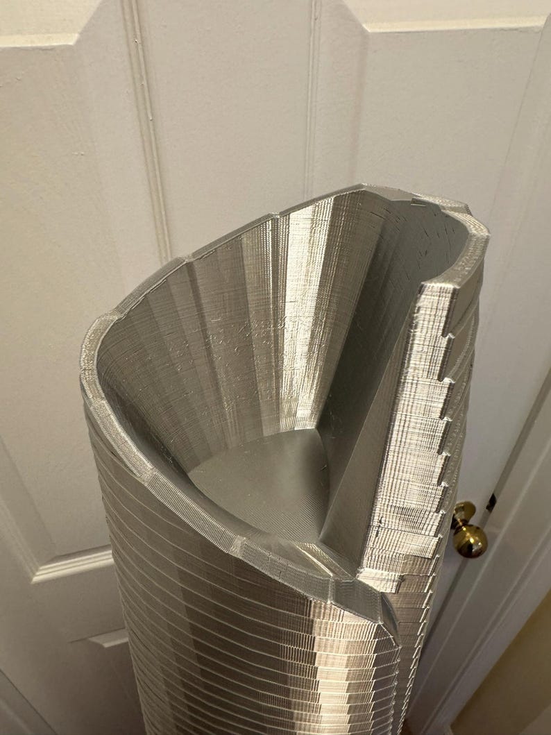May include: A silver 3D printed model of a tall, cylindrical building with a unique, angled design. The model is detailed with intricate lines and textures, showcasing the capabilities of 3D printing.