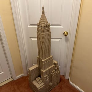 Peut inclure: Une grande maquette architecturale de couleur beige représentant un gratte-ciel, dotée d'une conception à plusieurs niveaux et d'une flèche pointue au sommet. La structure présente une grille de petites fenêtres en retrait sur toute sa façade.