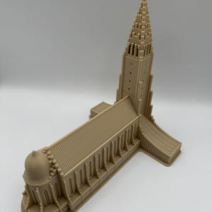 May include: A beige architectural model of a church with a tall, pointed steeple and a cross at the top. The building has multiple rectangular windows and a rounded dome. The model is made of a solid material and sits on a white surface.