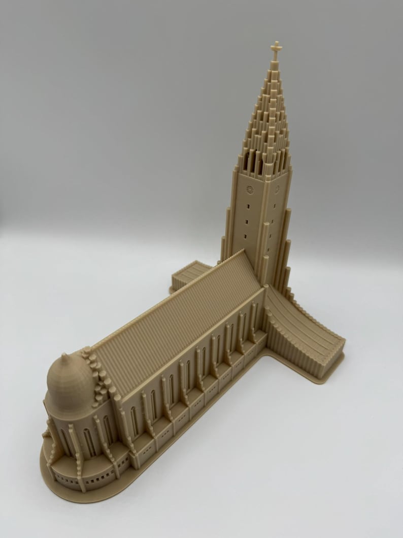 May include: A beige architectural model of a church with a tall, pointed steeple and a cross at the top. The building has multiple rectangular windows and a rounded dome. The model is made of a solid material and sits on a white surface.
