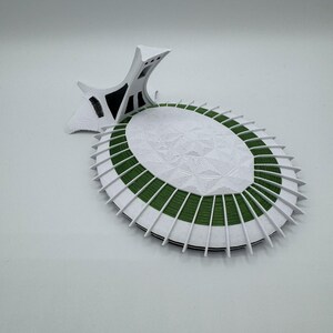 Puede incluir: Maqueta arquitect&oacute;nica blanca y verde con un dise&ntilde;o circular detallado. El modelo presenta una superficie central blanca texturizada rodeada de bandas verdes y blancas, y una estructura de soporte estilizada.