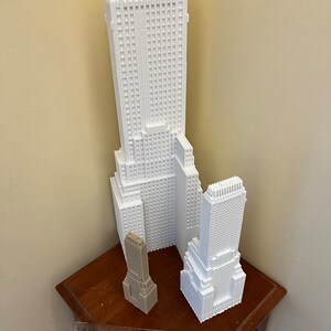 May include: Three white 3D printed models of skyscrapers. The tallest building is in the center, with a smaller building to the right and a very small building to the left. The buildings are on a wooden table with a ruler.