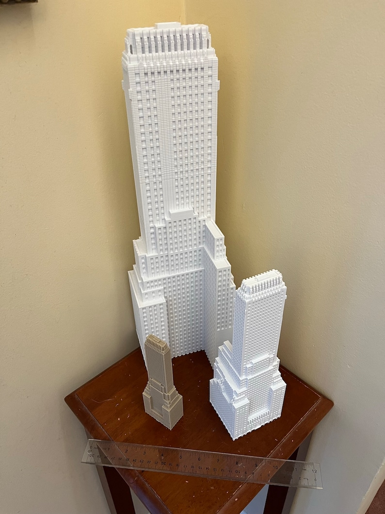 May include: Three white 3D printed models of skyscrapers. The tallest building is in the center, with a smaller building to the right and a very small building to the left. The buildings are on a wooden table with a ruler.