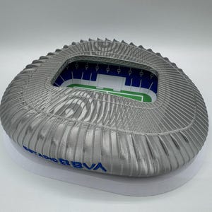 May include: A detailed model of a stadium, featuring a silver-colored exterior and a white base. The interior shows a green field and blue seating. The text "ESTADIO BBVA" is visible in blue.