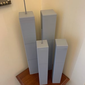 May include: Three grey model buildings, resembling skyscrapers, are arranged in a row on a wooden surface. The tallest building is on the left, with a smaller building to its right, and the shortest building is on the far right. A ruler is visible in the foreground.