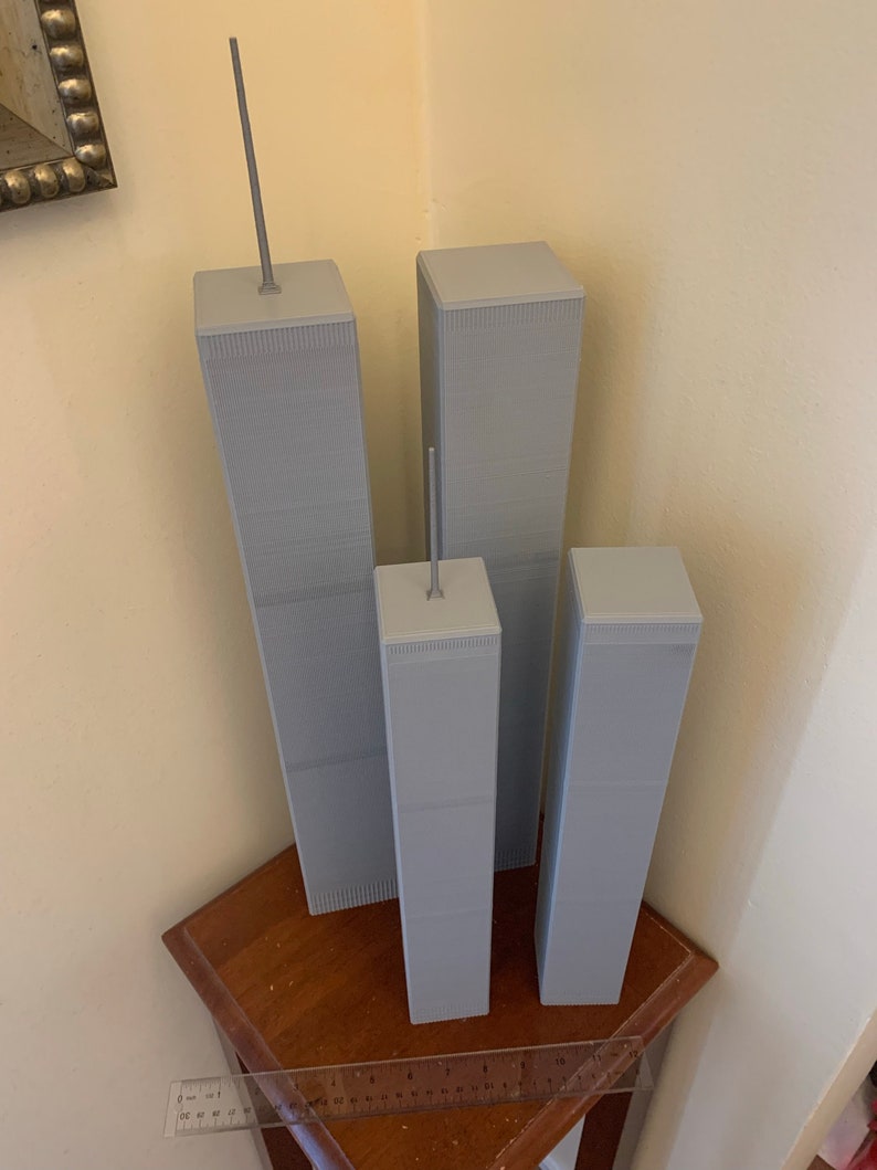 May include: Three grey model buildings, resembling skyscrapers, are arranged in a row on a wooden surface. The tallest building is on the left, with a smaller building to its right, and the shortest building is on the far right. A ruler is visible in the foreground.