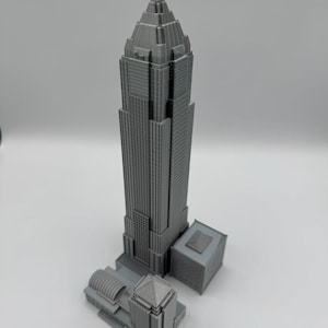 May include: A gray 3D-printed model of a skyscraper with a pointed top and multiple rectangular sections. The model includes a smaller building attached to the base, and is set against a white background.