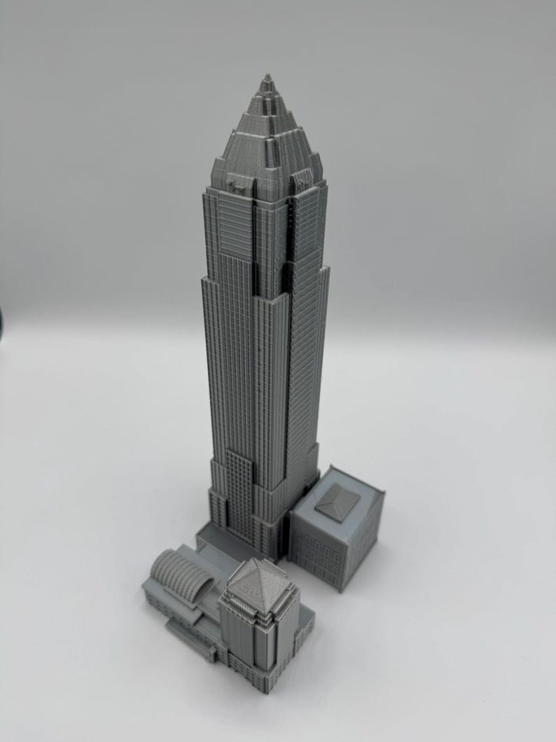 May include: A gray 3D-printed model of a skyscraper with a pointed top and multiple rectangular sections. The model includes a smaller building attached to the base, and is set against a white background.