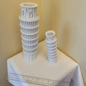 May include: Two white 3D printed models of the Leaning Tower of Pisa, one larger than the other, on a white tablecloth with a ruler measuring 12 inches.
