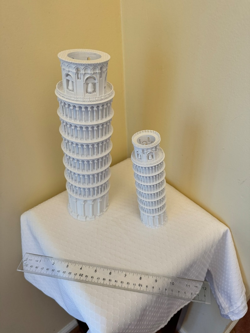 May include: Two white 3D printed models of the Leaning Tower of Pisa, one larger than the other, on a white tablecloth with a ruler measuring 12 inches.