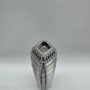May include: A silver-colored, geometric vase with a unique architectural design. The vase has a square top and a tapered body with layered, textured details. The overall aesthetic is modern and sculptural, suitable for home decor.