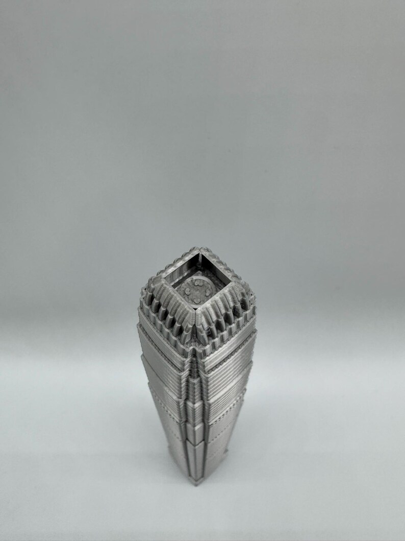 May include: A silver-colored, geometric vase with a unique architectural design. The vase has a square top and a tapered body with layered, textured details. The overall aesthetic is modern and sculptural, suitable for home decor.