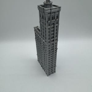 May include: A grey, 3D-printed model of a tall skyscraper. The building has many windows and architectural details, including a decorative top. The model is standing upright against a plain background.