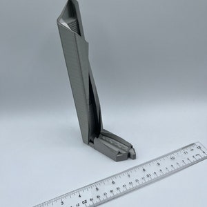 May include: A 3D printed model of a skyscraper, approximately 6 inches tall, with a gray finish. The model is sitting on a white surface with a ruler next to it.