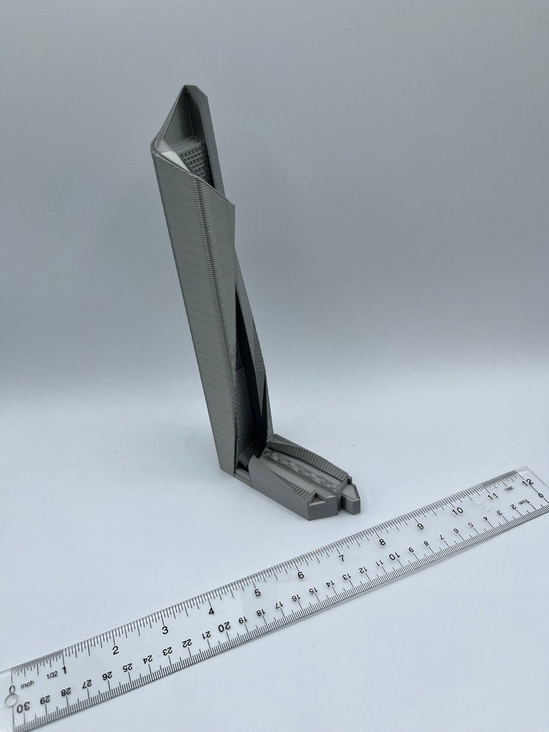 May include: A 3D printed model of a skyscraper, approximately 6 inches tall, with a gray finish. The model is sitting on a white surface with a ruler next to it.