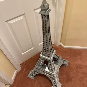 May include: A gray Eiffel Tower replica, standing tall on a brown carpet. The model features intricate lattice work and a detailed top spire. The tower is a decorative item, showcasing architectural design.