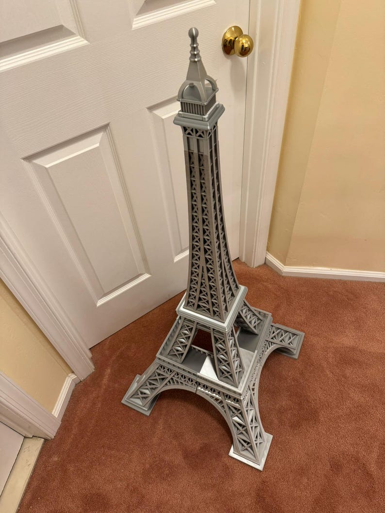 May include: A gray Eiffel Tower replica, standing tall on a brown carpet. The model features intricate lattice work and a detailed top spire. The tower is a decorative item, showcasing architectural design.