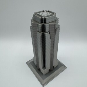 May include: A gray, 3D-printed model of a skyscraper. The building has a rectangular base and a tiered design, with a flat top featuring a circular indentation. The model is a solid gray color and sits on a white surface.