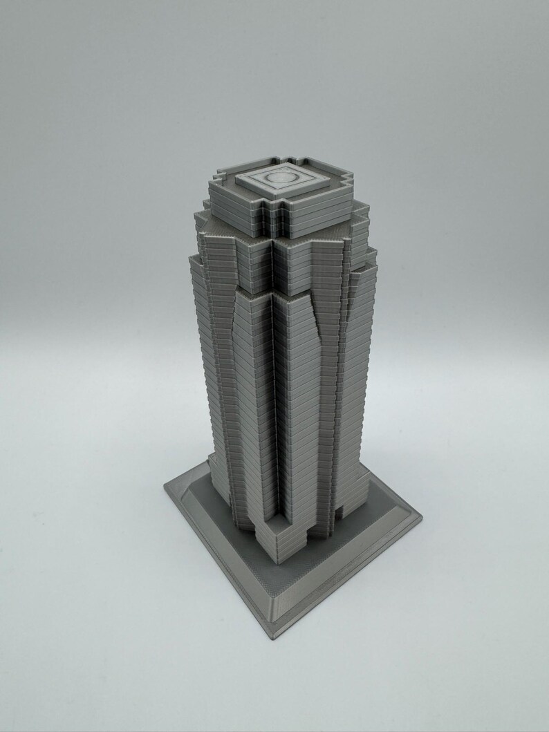 May include: A gray, 3D-printed model of a skyscraper. The building has a rectangular base and a tiered design, with a flat top featuring a circular indentation. The model is a solid gray color and sits on a white surface.