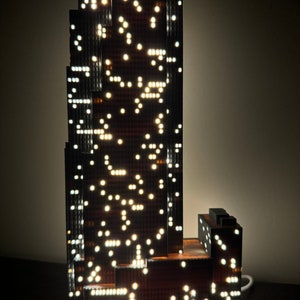 May include: A black and brown skyscraper model with white lights illuminating the windows. The model is lit from within and has a cord attached to the base.